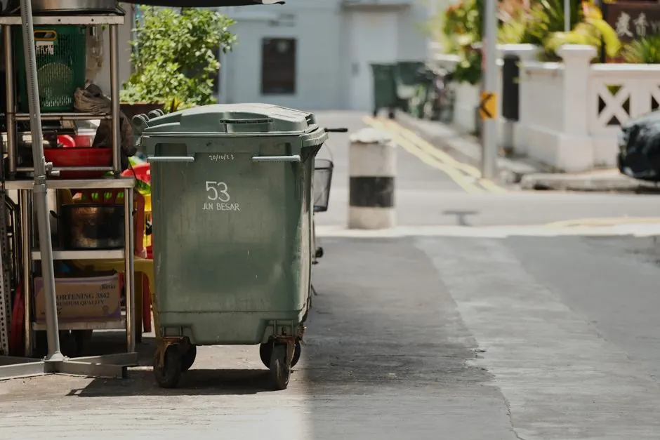 A green garbage bin on a quiet urban street under the sunlight.
