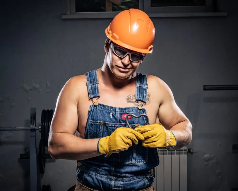 Muscular construction worker in overalls and helmet displaying strength and humor indoors.